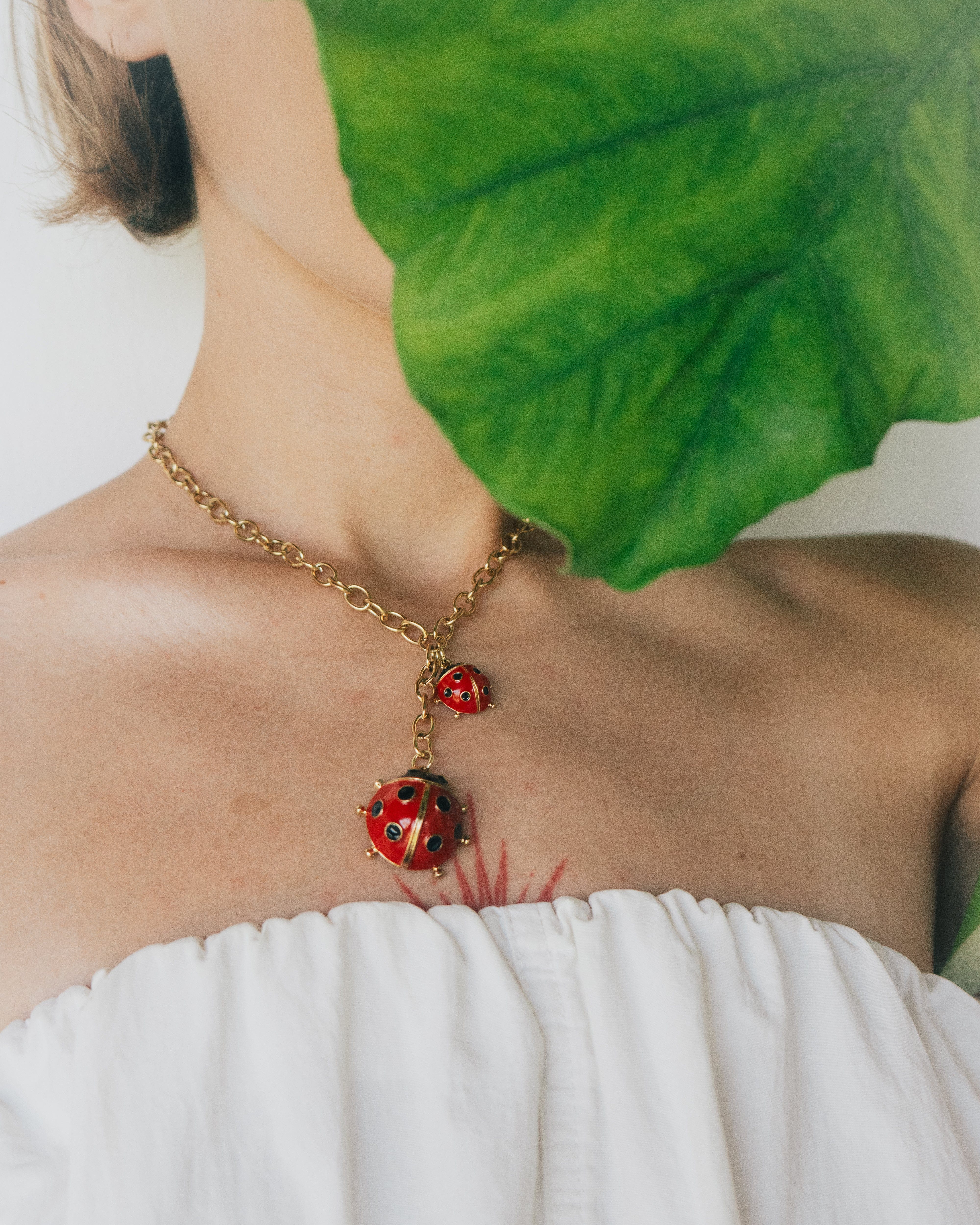 Person wearing a gold necklace with red ladybug pendants, partially covered by a green leaf.